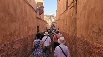 Tourists in hats fill a narrow, congested terracotta alleyway in the Marrakesh medina, Morocco, representing over-tourism.